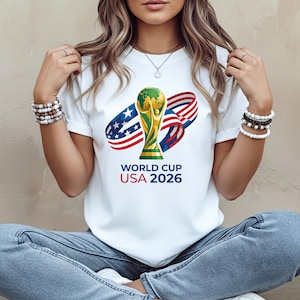 May include: White t-shirt featuring a graphic of the World Cup trophy with a green globe and gold accents, along with the text "WORLD CUP USA 2026". The design includes red, white, and blue ribbons.