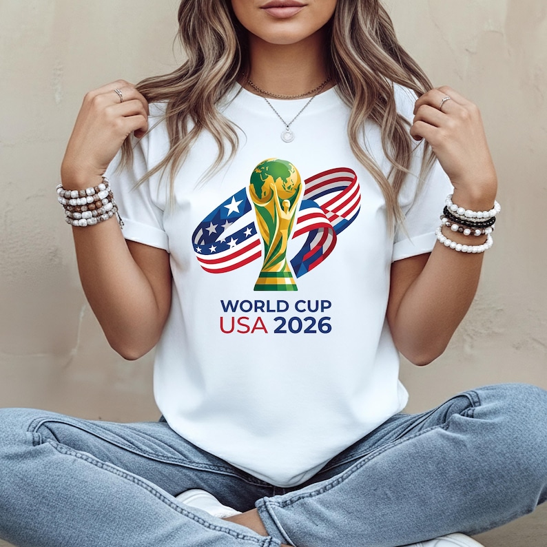 May include: White t-shirt featuring a graphic of the World Cup trophy with a green globe and gold accents, along with the text "WORLD CUP USA 2026". The design includes red, white, and blue ribbons.