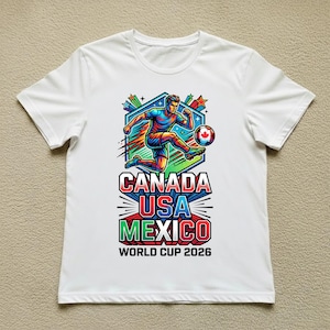 May include: White t-shirt featuring a colorful graphic of a soccer player kicking a ball. The design includes the text "CANADA USA MEXICO WORLD CUP 2026" in bold, vibrant colors. The shirt is a casual wear item.