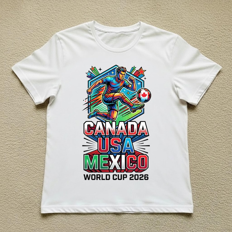 May include: White t-shirt featuring a colorful graphic of a soccer player kicking a ball. The design includes the text "CANADA USA MEXICO WORLD CUP 2026" in bold, vibrant colors. The shirt is a casual wear item.