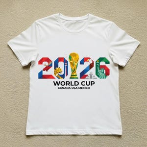 May include: White t-shirt featuring the text "2026 WORLD CUP CANADA USA MEXICO" in colorful block letters. The design includes a trophy, a moose, a jaguar, an eagle, and the Statue of Liberty.