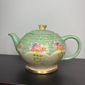 Könnte beinhalten: Vintage Teekanne mit rundem Korpus, hellgrüner Farbe und floralen Designs in Rosa, Weiß und Gelb. Deckel und Sockel sind mit goldenen Akzenten versehen. Die Teekanne hat einen gebogenen Griff und Ausguss.