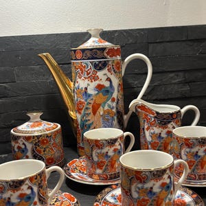 May include: A complete porcelain tea set with a peacock design. The set includes a tall teapot with a gold spout, a creamer, a sugar bowl, six teacups, and saucers. The cups and saucers are decorated with a vibrant peacock and floral pattern.