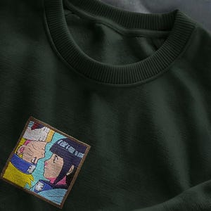 Può includere: Felpa verde scuro con girocollo e una piccola toppa ricamata. La toppa presenta un'illustrazione colorata in stile anime di due personaggi che si guardano. La felpa è realizzata in un tessuto morbido e strutturato.