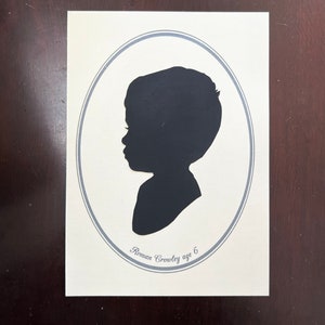 May include: A black silhouette of a child's profile, set within a gray oval frame on an off-white background. The text "Roman Crowley age 6" is printed below the silhouette. The artwork is mounted on a rectangular surface.