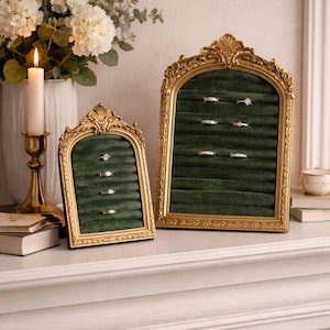 May include: Two ornate gold ring displays with green velvet interiors. Each display holds several rings. The displays are on a white surface, with a candle, flowers, and books in the background. The displays have an arched shape.