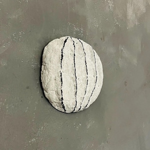 May include: A white, oval-shaped sculpture with vertical black stripes. The artwork is set against a gray background, creating a contrast between the textured sculpture and the smooth surface.