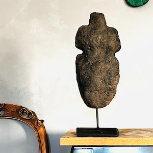 Puede incluir: Una escultura de piedra marrón desgastada con forma humana, montada sobre un soporte de metal negro. La escultura tiene una superficie rugosa y texturizada y está sobre una mesa de madera. El fondo es una pared gris claro.