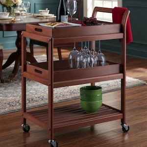 May include: A brown wooden rolling bar cart with three tiers, featuring a top tray with a wine bottle, a plate of snacks, and grapes. The middle tier holds hanging wine glasses, and the bottom tier holds three stacked green bowls. A red towel hangs from the side handle.