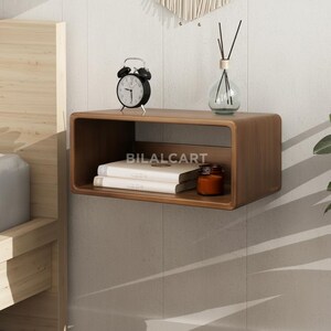 Puede incluir: Estanter&iacute;a flotante de madera rectangular con esquinas redondeadas. Un despertador negro, libros y un tarro peque&ntilde;o est&aacute;n sobre ella. Tambi&eacute;n se ven un difusor de vidrio y decoraci&oacute;n de pared. El texto "BILALCART" est&aacute; impreso en la estanter&iacute;a.