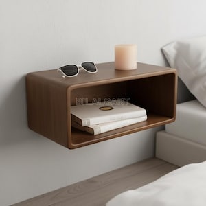 May include: A dark brown, floating wooden bedside table with rounded edges, mounted on a white wall. It features sunglasses, a beige candle, and two books. The rectangular table has an open front. The text "BILALCART" is visible. The unit is designed for a bedroom.