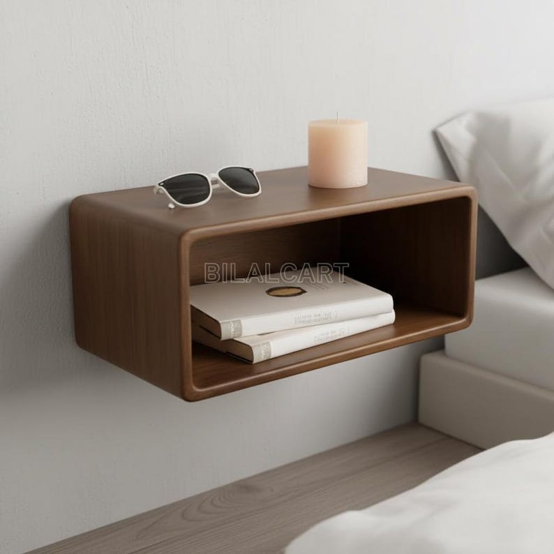 Puede incluir: Una mesita de noche flotante de madera marr&oacute;n oscuro con bordes redondeados, montada en una pared blanca. Contiene gafas de sol, una vela beige y dos libros. La mesa rectangular tiene un frente abierto. El texto "BILALCART" es visible.