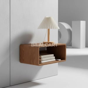 Puede incluir: Una mesita de noche flotante de madera, con un acabado natural. Tiene un dise&ntilde;o rectangular con bordes redondeados y una balda inferior con libros y un mando a distancia. Una peque&ntilde;a l&aacute;mpara con pantalla plisada est&aacute; encima. El texto "BILALCART" es visible.