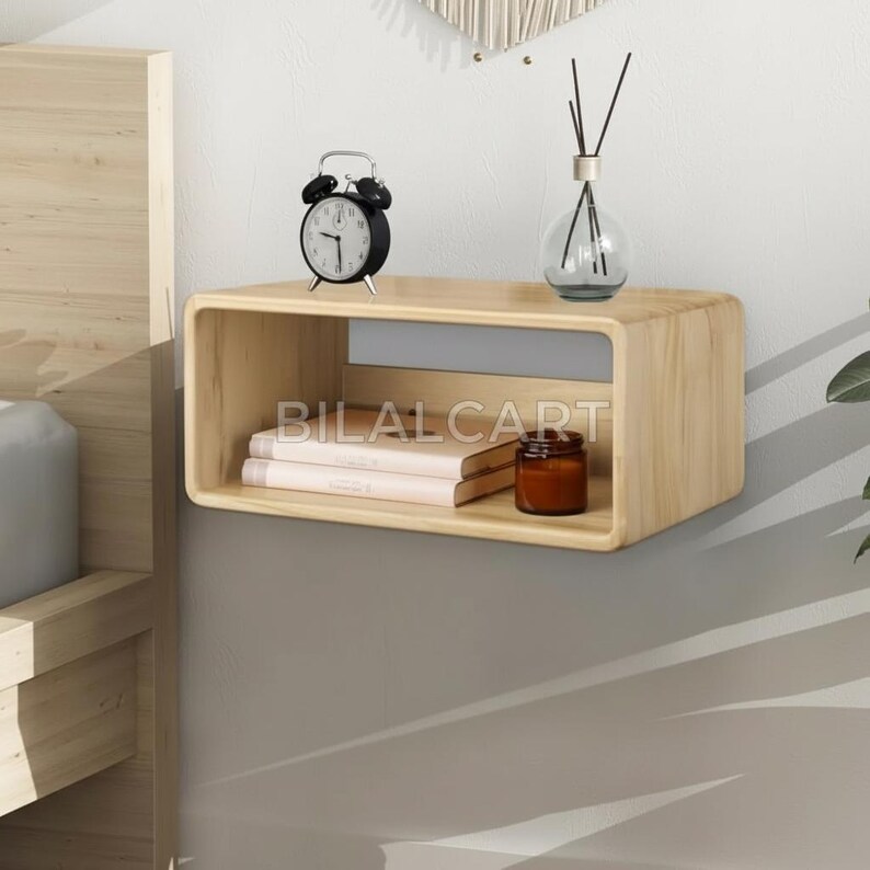 Puede incluir: Una estanter&iacute;a flotante de madera clara con esquinas redondeadas, que sostiene un despertador negro, dos libros, un peque&ntilde;o tarro marr&oacute;n y un difusor de vidrio. La estanter&iacute;a est&aacute; montada en una pared de dos tonos. El texto "BILALCART" es visible.