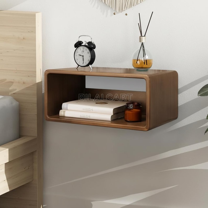 Puede incluir: Una mesita de noche rectangular de madera, flotante, con bordes redondeados. Contiene un despertador negro, dos libros, un peque&ntilde;o tarro y un difusor de vidrio. El texto "BILALCART" es visible en la parte frontal.