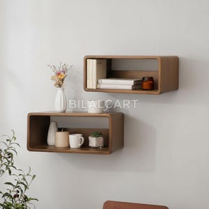 Puede incluir: Dos estanter&iacute;as flotantes rectangulares de madera montadas en una pared blanca. Las estanter&iacute;as est&aacute;n llenas de objetos decorativos, incluyendo libros, velas, una peque&ntilde;a planta y un jarr&oacute;n con flores. Las estanter&iacute;as son de color marr&oacute;n c&aacute;lido.