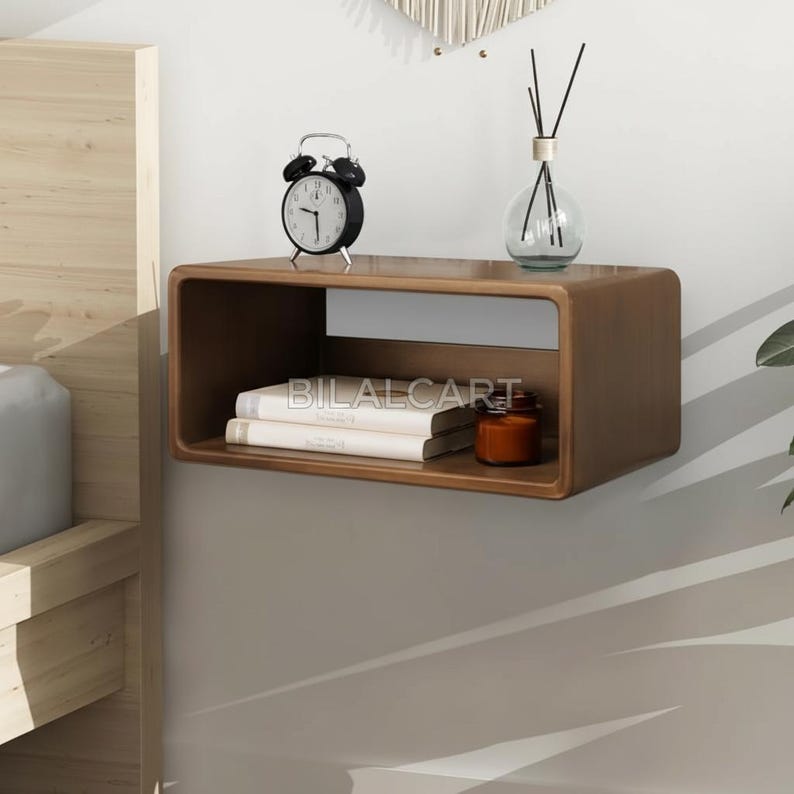 Puede incluir: Una mesita de noche flotante marr&oacute;n con un dise&ntilde;o rectangular. Un despertador negro, un difusor de vidrio, libros y una peque&ntilde;a vela naranja est&aacute;n sobre la mesita. El texto "BILALCART" es visible.