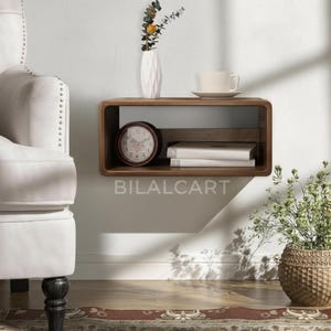 Puede incluir: Estante flotante de madera con bordes redondeados, con un jarr&oacute;n blanco con flores, una taza de t&eacute;, libros y un reloj. El estante est&aacute; montado en una pared blanca, con una vista parcial de una silla y una cesta de mimbre con vegetaci&oacute;n.