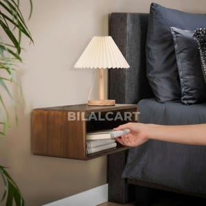 Puede incluir: Una mesita de noche flotante de madera oscura con un borde frontal curvado est&aacute; montada en una pared beige. Una peque&ntilde;a pantalla de l&aacute;mpara plisada de color crema se encuentra encima de la mesita de noche, junto a una pila de libros. El texto "BILALCART" est&aacute; impreso en la mesita de noche.