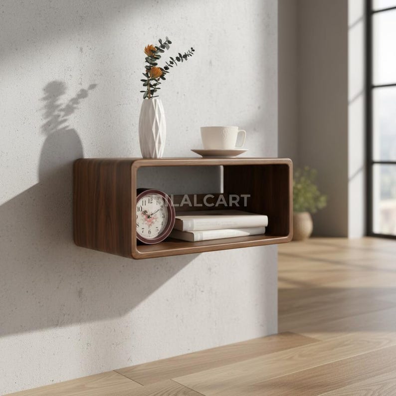 Puede incluir: Estanter&iacute;a rectangular de madera, montada en la pared, con bordes redondeados. Muestra un jarr&oacute;n blanco con flores, una taza y platillo, un reloj y libros. El texto "LALCART" es visible en la estanter&iacute;a.