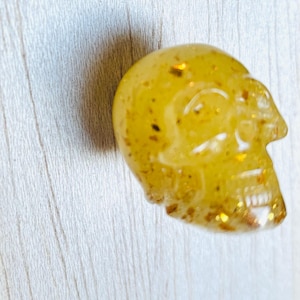 May include: A small, yellow skull-shaped object with gold glitter embedded throughout. The skull is detailed with visible eye sockets, a nose cavity, and teeth. The object is likely made of resin or a similar material and is set against a light wood grain background.