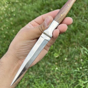 Puede incluir: Un cuchillo de hoja fija con una hoja de acero plateada, afilada y puntiaguda, y un mango de madera marrón lisa asegurado con tres pequeños pasadores de latón. El refuerzo de metal conecta la hoja con el mango, creando un diseño recto.