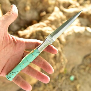 Op de afbeelding: Een puntige metalen briefopener met een cilindervormig handvat in een lichtturquoise afwerking, geaccentueerd door donkerbruine aders en kleine goudkleurige ronde inlegstukken. Het lemmet is glad, zilverkleurig en loopt spits toe.
