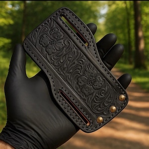 Puede incluir: Funda de cuero negro con herramientas florales. La funda presenta un patrón floral en relieve detallado, bordes cosidos y remaches de latón. El diseño incluye dos presillas para cinturón y un interior rojo contrastante.