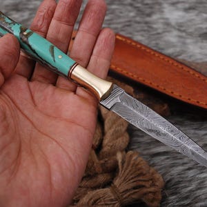May include: A fixed-blade knife featuring a pointed Damascus steel blade with a distinct wavy pattern, a polished brass bolster, and a handle crafted from turquoise-coloured material with dark veining, accompanied by a tan leather sheath.