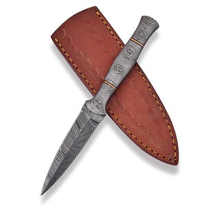 May include: A Damascus steel dagger with a patterned blade and handle, featuring gold accents. The dagger rests on a brown leather sheath with yellow stitching. The blade is sharp and pointed, and the handle has a unique swirl design.