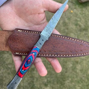 May include: A fixed-blade knife featuring a pointed Damascus steel blade with a distinct wavy pattern. The handle is crafted from layered red and blue material secured with two metal pins. It is accompanied by a brown leather sheath with stitching.