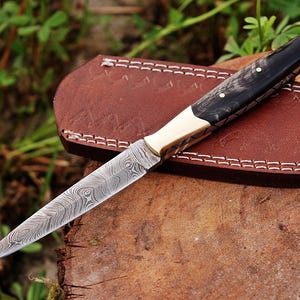 May include: A fixed-blade knife with a patterned Damascus steel blade, a brass bolster, and a dark, polished handle secured with two pins. The knife is accompanied by a brown leather sheath featuring white stitching and the number 1616.