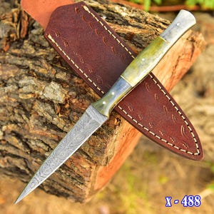 Puede incluir: Un cuchillo de hoja fija con una hoja puntiaguda de acero Damasco con patrón y un mango liso de color claro asegurado con tres pasadores de metal. El cuchillo viene acompañado de una funda de cuero marrón oscuro con costuras decorativas.