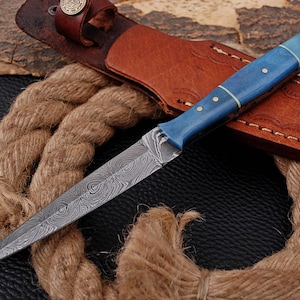 Op de afbeelding: Een mes met een vast lemmet en een puntig Damascus stalen blad met een wervelend patroon. Het handvat is felblauw met drie koperen pinnen. Het mes wordt vergezeld door een bruine lederen schede met een drukknoop en een dik touw.