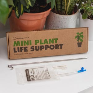 May include: A brown cardboard box with the text "MINI PLANT LIFE SUPPORT" in green and black. A metal rod and a small plastic bag with a clear tube and blue valve are in front of the box. The box has a plant illustration.