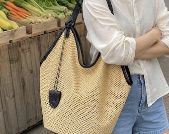 May include: A large hobo-style shoulder bag crafted from a woven tan straw-like material, featuring black leather trim along the edges and a matching black leather shoulder strap. A small black leather tag hangs from the top handle attachment.