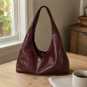 May include: A deep burgundy hobo bag with a smooth, slightly glossy finish. The bag features a single integrated shoulder strap, a slouchy crescent shape, and a central vertical seam running down the front panel, creating a clean, minimalist silhouette.