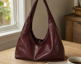 May include: A deep burgundy hobo bag with a smooth, slightly glossy finish. The bag features a single integrated shoulder strap, a slouchy crescent shape, and a central vertical seam running down the front panel, creating a clean, minimalist silhouette.