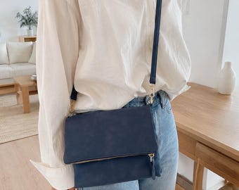 May include: A navy blue fold-over crossbody bag with a gold-tone zipper closure and a matching thin shoulder strap. The bag is worn over a white button-down shirt and light blue denim jeans, featuring a smooth texture and a rectangular shape.