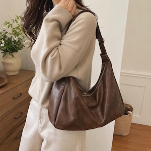 May include: A dark brown hobo-style shoulder bag with a smooth, slightly distressed finish. The bag features a slouchy, crescent-shaped silhouette, a single wide shoulder strap with an adjustable buckle, and a top zipper closure with a metal pull.