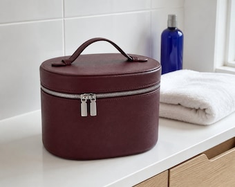 May include: A deep burgundy oval-shaped vanity case with a textured finish, a top handle, and a silver-toned double zipper closure. The structured container features a clean, minimalist design with a secure zip mechanism for storage.