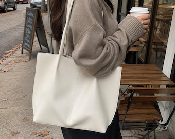 May include: A solid off-white tote bag with a smooth finish, featuring two long shoulder straps and a vertical seam detail running down the center of the front panel. The bag has a rectangular shape with a wide opening and a clean, simple design.