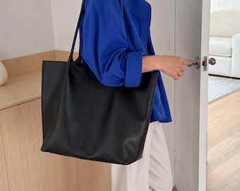 May include: A large, black, rectangular tote bag with a smooth, matte finish and two long, thin shoulder straps. The bag features a vertical seam running down the center of the front panel, providing a structured appearance for daily use.