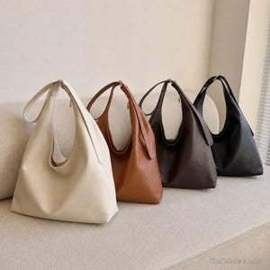 May include: Four hobo bags in a row, each featuring a single shoulder strap and a textured surface. The bags are arranged by color from left to right: cream, tan, dark brown, and black. Each bag has a slouchy, unstructured shape.