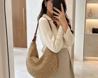 May include: A crescent-shaped hobo bag crafted from woven tan straw material. The bag features a single brown leather shoulder strap attached with metal hardware, a curved silhouette, and a textured exterior surface with a natural beige color.