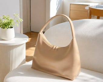May include: A smooth, beige hobo-style shoulder bag with a single arched handle and a top zipper closure featuring two matching zipper pulls. The bag has a curved, crescent-like silhouette and a clean, solid-colored exterior surface.