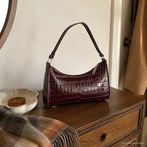 May include: A dark burgundy shoulder bag with a glossy crocodile-embossed texture. The bag features a curved top silhouette, a single thin handle attached with gold-tone hardware, and a gold-tone zipper closure along the top edge.