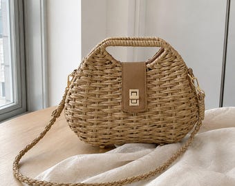May include: A woven straw handbag with a rounded silhouette and a tan faux leather center flap featuring a gold-tone metal turn-lock closure. The bag includes a braided straw handle and a detachable braided straw shoulder strap with gold hardware.