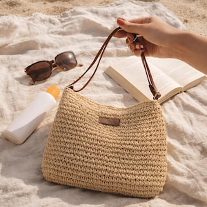 Puede incluir: Un bolso beige tejido con correas de cuero marrón y una pequeña etiqueta de cuero marrón. El bolso está sujeto por una mano. También se ven gafas de sol, protector solar y un libro.