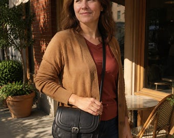 May include: A black leather crossbody bag with a rounded flap, a vertical strap detail, and a silver-toned metal buckle closure. The bag features a slim, adjustable shoulder strap and a textured surface, worn over a tan cardigan and a rust-colored top.